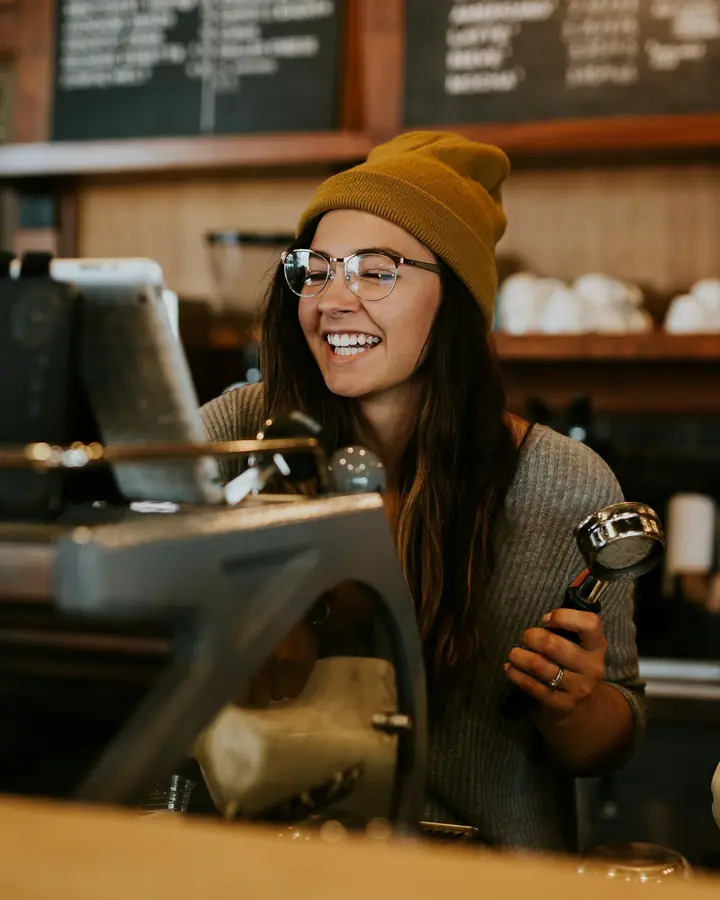 Een lachende barista met een muts achter de espressomachine